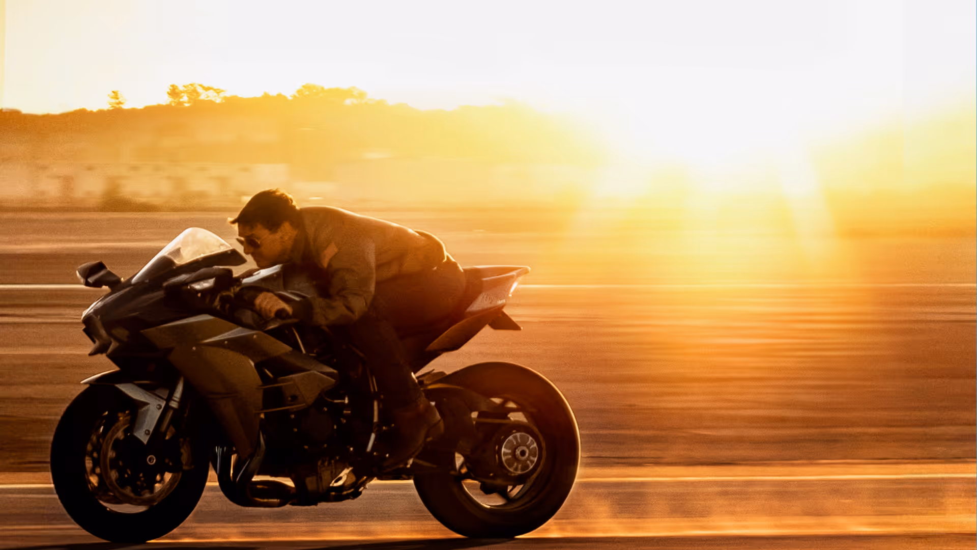 Person in sunglasses leaning forward riding a black motorcycle on a road at sunset.