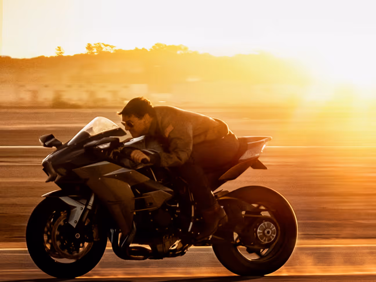 Person wearing sunglasses riding a motorcycle with sun setting in the background.