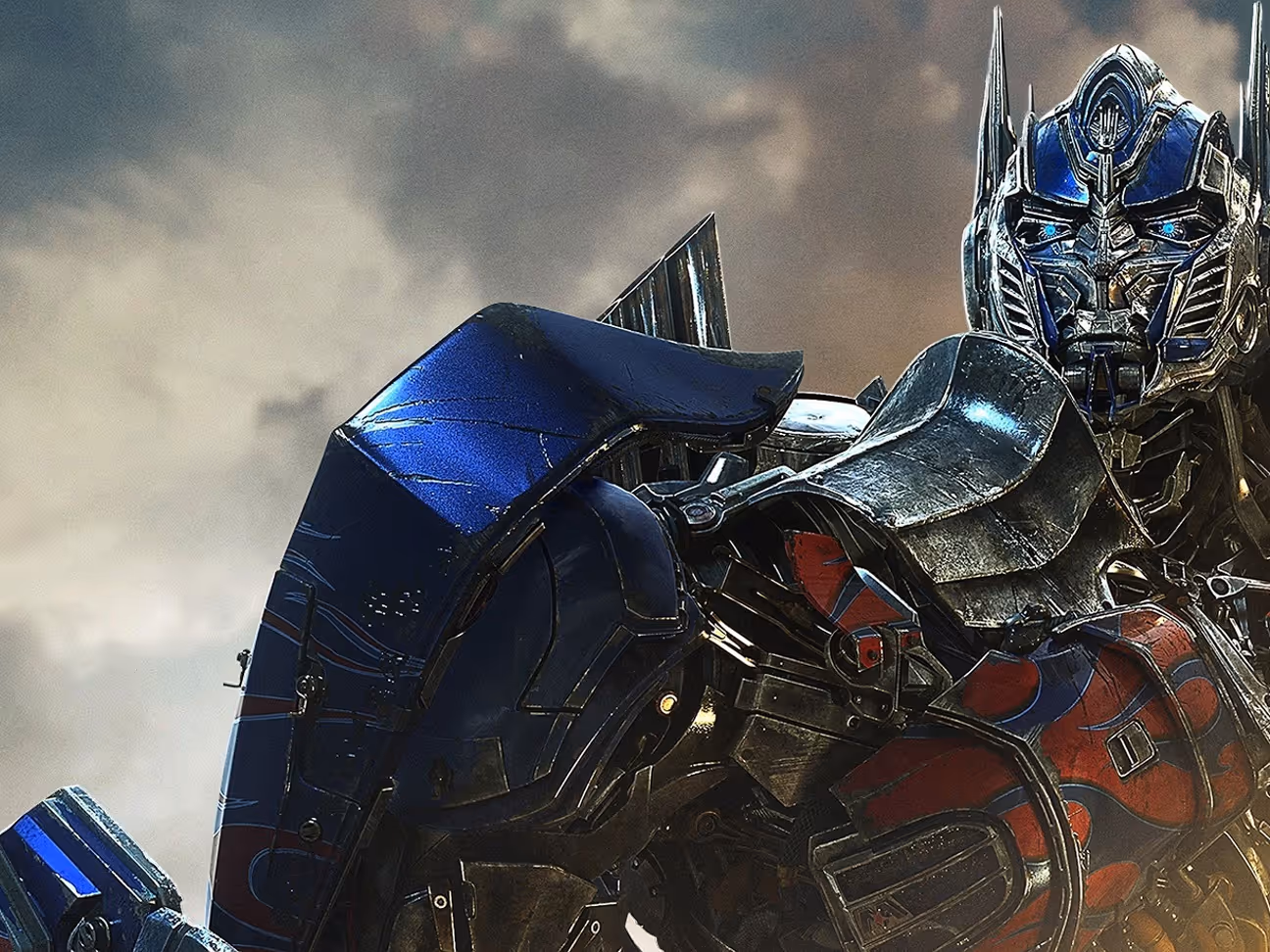 Close-up of a blue and red detailed robotic figure with glowing blue eyes against a cloudy sky.